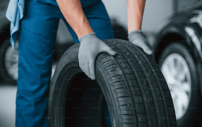 A mechanic in blue overalls and grey gloves holding a black car tire in a garage setting