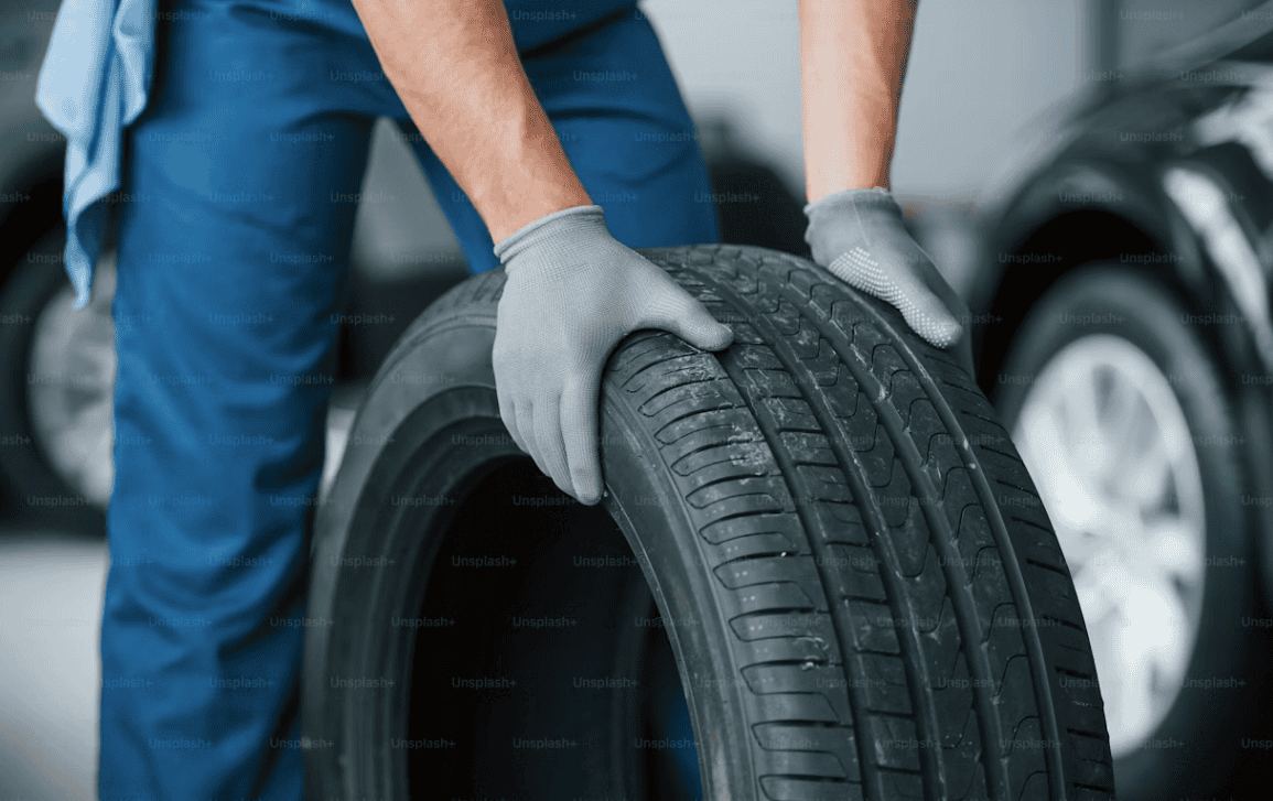 A mechanic in blue overalls and grey gloves holding a black car tire in a garage setting